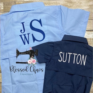 Puede incluir: Dos camisas de botones azules y azul marino con letras bordadas. La camisa azul claro tiene las letras "JWS" bordadas en la espalda. La camisa azul marino tiene la palabra "SUTTON" bordada en la espalda. Ambas camisas son de manga corta y tienen un bolsillo en el lado izquierdo.