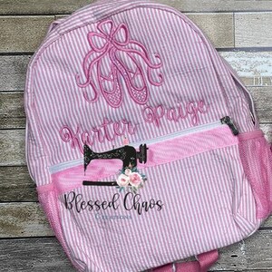 May include: Pink and white striped backpack with a pink ballerina shoe design and the name "Karter Paige" embroidered on the front. The backpack also has a pink ribbon with the text "Blessed Chaos Creations" embroidered on it.