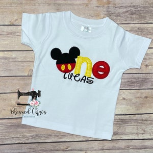 Peut inclure: T-shirt &agrave; manches courtes blanche avec un motif Mickey Mouse rouge, jaune et noir et le mot "one" en jaune et rouge. Le nom "Lucas" est brod&eacute; en jaune sous le motif.