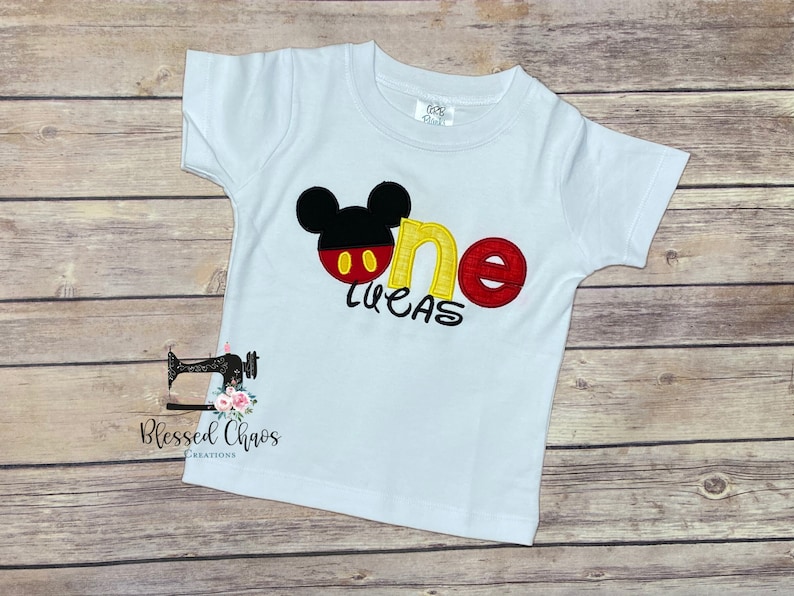 Peut inclure: T-shirt &agrave; manches courtes blanche avec un motif Mickey Mouse rouge, jaune et noir et le mot "one" en jaune et rouge. Le nom "Lucas" est brod&eacute; en jaune sous le motif.