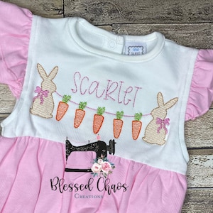 May include: A pink and white baby onesie with a bunny and carrot garland, the name "Scarlett" embroidered on the front, and the text "Blessed Chaos Creations" on the bottom.