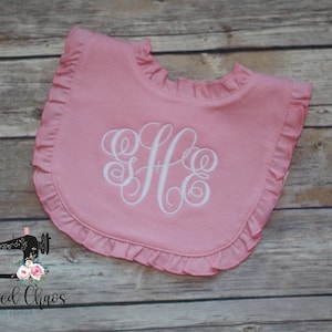 May include: Pink ruffled baby bib with a white embroidered monogram "S.H.E."