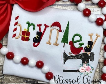 May include: White Christmas-themed shirt with the name "Brynnley" embroidered in green, red, and brown. The letters are decorated with Christmas-themed embellishments, including a candy cane, a gift box, a Santa hat, a Christmas tree, and a boot. A black sewing machine with pink flowers is embroidered below the name. The text "Blessed Chaos Creations" is embroidered in black at the bottom of the shirt.