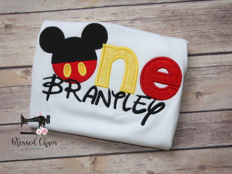 Peut inclure: Combinaison blanche avec un motif Mickey Mouse brod&eacute; en rouge, jaune et noir. La combinaison dit "one" en jaune et rouge avec le nom "Branpley" en noir.
