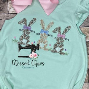 Könnte beinhalten: Hellblaues Baby-Body mit gestickten Hasen mit Schleifen, einer Nähmaschine und dem Text "Blessed Chaos Creations". Oben befindet sich eine große rosa Schleife.