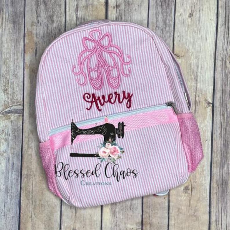 May include: Pink and white striped backpack with a pink ballet shoe design and the name "Avery" embroidered on the front. The backpack has a pink zippered pocket on the front and a mesh side pocket. The bottom of the backpack has a pink and white striped fabric with the words "Blessed Chaos Creations" embroidered on it.