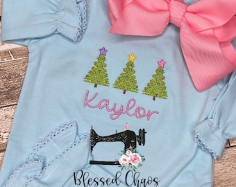May include: Light blue baby onesie with embroidered Christmas trees, the name "Kaylor", a sewing machine, and the text "Blessed Chaos Creations".