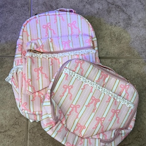 Pode incluir: Uma mochila e uma bolsa quadrada a condizer com riscas cor-de-rosa e brancas, ambas com um padrão repetitivo de laços cor-de-rosa e detalhes em glitter dourado. Cada item inclui um bolso frontal com fecho, folho branco e ferragens douradas.