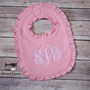 May include: A pink baby bib with a white ruffled edge and a white monogram of the letters "SPS" embroidered in the center.