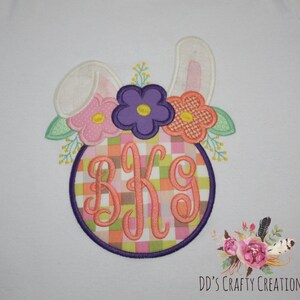 Puede incluir: Tela blanca con un dise&ntilde;o de conejo de Pascua bordado. El conejo tiene orejas rosas y sostiene un c&iacute;rculo con un patr&oacute;n de cuadros rosa, verde y naranja. El c&iacute;rculo tiene las letras "BKG" bordadas en naranja. El conejo est&aacute; rodeado de flores rosas, moradas y naranjas.