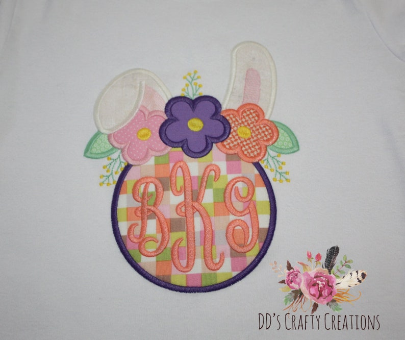 Puede incluir: Tela blanca con un dise&ntilde;o de conejo de Pascua bordado. El conejo tiene orejas rosas y sostiene un c&iacute;rculo con un patr&oacute;n de cuadros rosa, verde y naranja. El c&iacute;rculo tiene las letras "BKG" bordadas en naranja. El conejo est&aacute; rodeado de flores rosas, moradas y naranjas.
