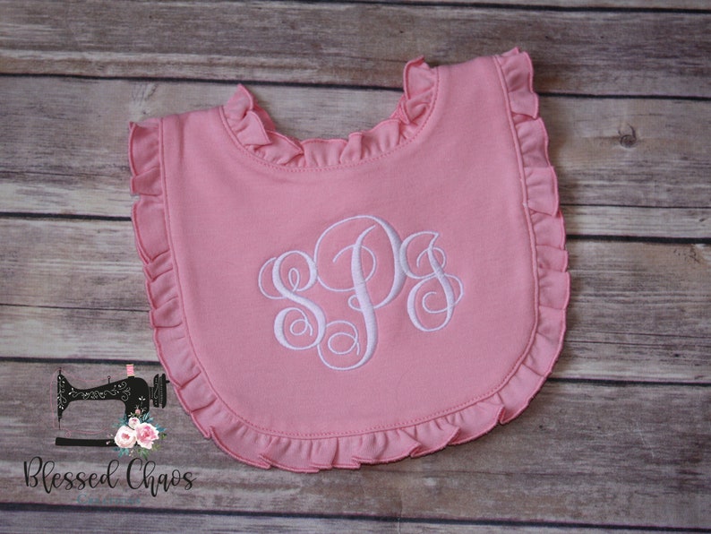 May include: Pink baby bib with a white ruffled edge and a white embroidered monogram "SPS".