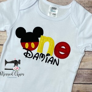 Peut inclure: Un body b&eacute;b&eacute; blanc avec une t&ecirc;te de Mickey Mouse noire, un 'un' rouge et jaune et le nom 'Damian' brod&eacute; en jaune. Le body est de taille 12-18 mois.