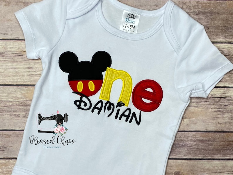 Peut inclure: Un body b&eacute;b&eacute; blanc avec une t&ecirc;te de Mickey Mouse noire, un 'un' rouge et jaune et le nom 'Damian' brod&eacute; en jaune. Le body est de taille 12-18 mois.
