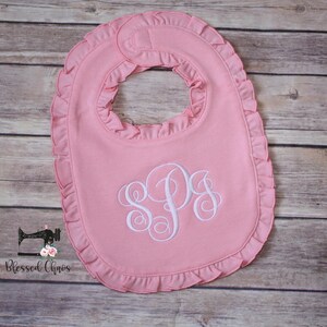 May include: A pink baby bib with a white ruffled edge and a white monogram of the letters "SPS" embroidered in the center.