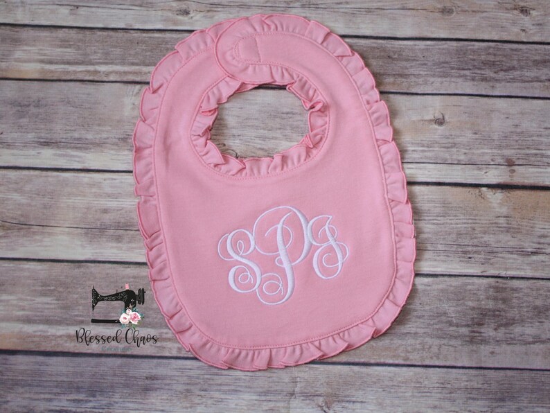 May include: A pink baby bib with a white ruffled edge and a white monogram of the letters "SPS" embroidered in the center.