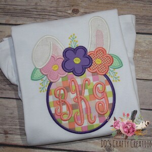 Puede incluir: Camiseta blanca con un dise&ntilde;o bordado de un conejito con orejas rosas, flores y un monograma en un c&iacute;rculo con un patr&oacute;n de cuadros. El monograma es "BKG".