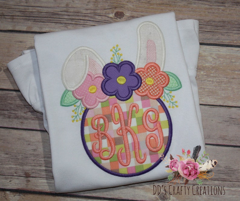 Puede incluir: Camiseta blanca con un dise&ntilde;o bordado de un conejito con orejas rosas, flores y un monograma en un c&iacute;rculo con un patr&oacute;n de cuadros. El monograma es "BKG".