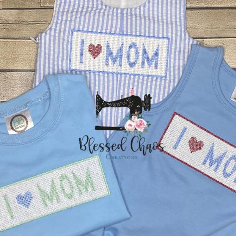 May include: Three blue shirts with embroidered "I love Mom" designs. The shirts are in different shades of blue and have different colored hearts. The designs are in a cross-stitch style.