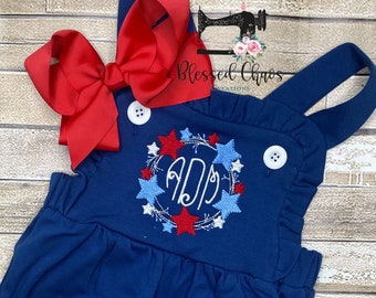 May include: Blue denim overalls with a red, white, and blue star and firework wreath with a monogram in the center. The overalls have a ruffled neckline and adjustable straps.