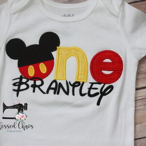 Peut inclure: Combinaison blanche avec une t&ecirc;te de Mickey Mouse noire, un 'un' rouge et jaune et le nom 'BRANPLY' en noir. La combinaison a un petit logo de machine &agrave; coudre avec le texte 'Blessed Chaos Creations'.