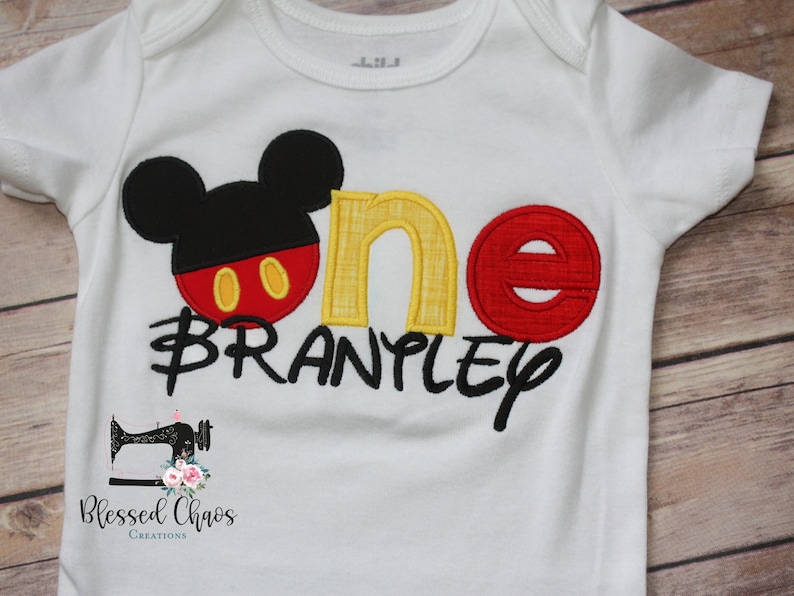 Peut inclure: Combinaison blanche avec une t&ecirc;te de Mickey Mouse noire, un 'un' rouge et jaune et le nom 'BRANPLY' en noir. La combinaison a un petit logo de machine &agrave; coudre avec le texte 'Blessed Chaos Creations'.