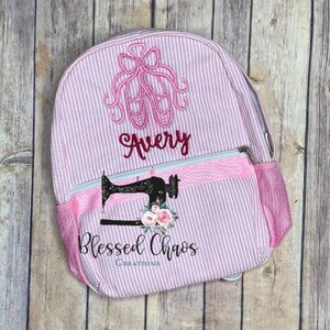 May include: Pink and white striped backpack with a pink ballet shoe design and the name Avery embroidered on the front. The backpack has a pink zippered pocket on the front and a mesh side pocket. The bottom of the backpack has a pink and white striped fabric with the words "Blessed Chaos Creations" embroidered on it.