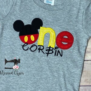 Peut inclure: T-shirt &agrave; manches courtes gris avec un motif Mickey Mouse brod&eacute; en rouge, jaune et noir. Le t-shirt dit "one" en jaune et rouge avec le nom "Corbin" en dessous.
