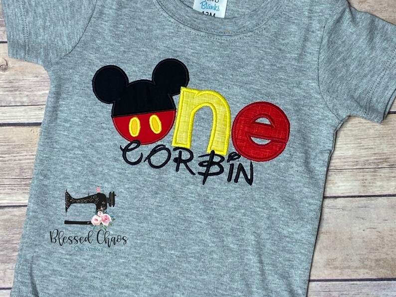 Peut inclure: T-shirt &agrave; manches courtes gris avec un motif Mickey Mouse brod&eacute; en rouge, jaune et noir. Le t-shirt dit "one" en jaune et rouge avec le nom "Corbin" en dessous.