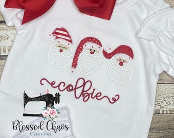 May include: White onesie with red and white embroidered Santa faces and the text "colbie" in red. The onesie has a red bow on the shoulder and a white lace trim around the neckline.