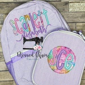 May include: Purple and white striped backpack and lunch bag set with embroidered names "Scarlett Elizabeth" and "Sc" in colourful letters. The backpack has a mesh pocket and the lunch bag has a zipper closure. The set is from Blessed Chaos Creations.