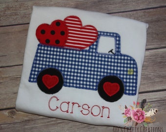 May include: A white t-shirt with a blue and white gingham truck carrying red and black hearts. The name "Carson" is embroidered on the shirt.