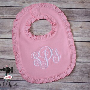 May include: Pink baby bib with a white ruffled edge and a white monogram of the letters "SPS".