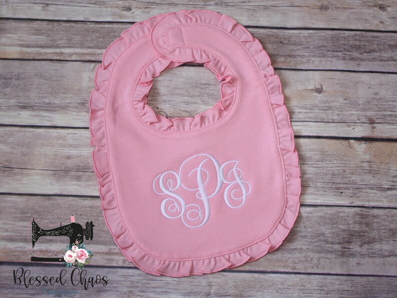 May include: Pink baby bib with a white ruffled edge and a white monogram of the letters "SPS".
