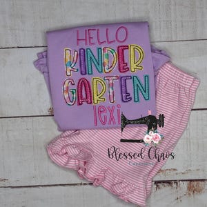 May include: A lavender long-sleeve shirt and pink striped pants set. The shirt has the words "Hello Kindergarten Lexi" embroidered in colorful letters. The pants have a ruffled hem. The set is from Blessed Chaos Creations.