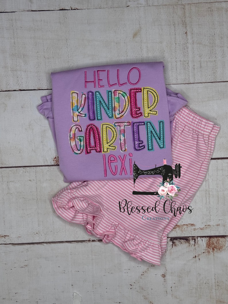 May include: A lavender long-sleeve shirt and pink striped pants set. The shirt has the words "Hello Kindergarten Lexi" embroidered in colorful letters. The pants have a ruffled hem. The set is from Blessed Chaos Creations.