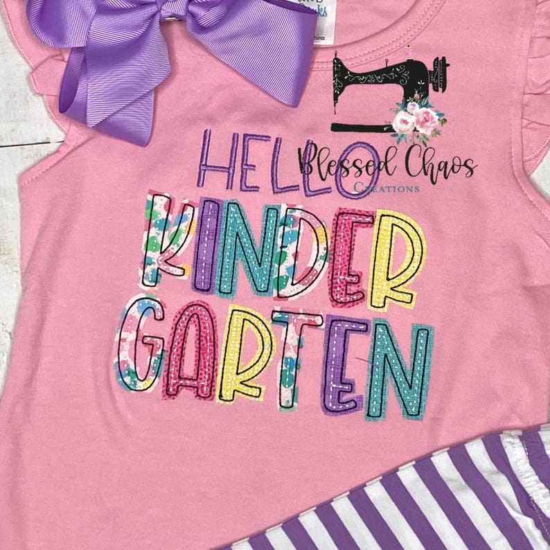 May include: Pink sleeveless top with the words "Hello Kindergarten" in colorful embroidered letters. A purple bow is attached at the top. The top has a sewing machine graphic and the words "Blessed Chaos Creations".