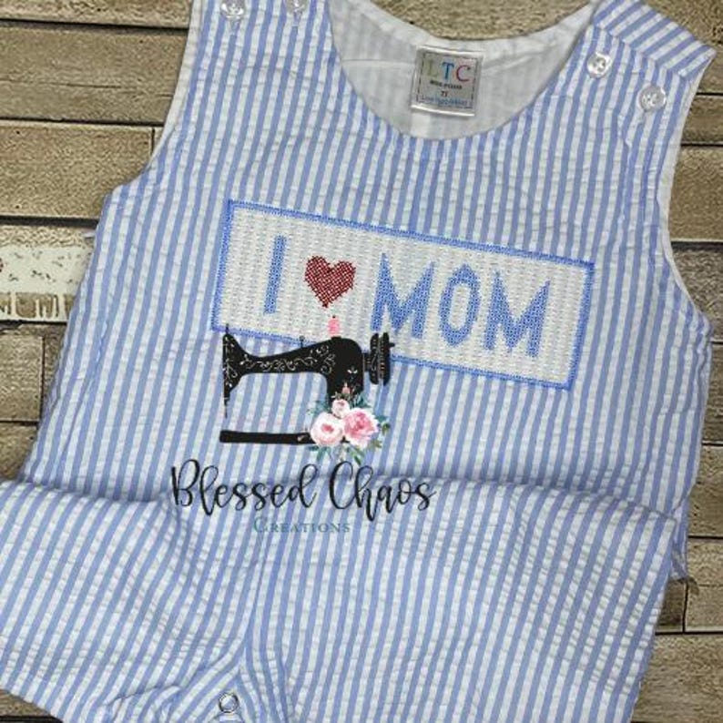 May include: A blue and white striped baby onesie with a white rectangle that says "I love MOM" in red and white embroidery. A black sewing machine with pink flowers is below the rectangle. The text "Blessed Chaos Creations" is below the sewing machine.