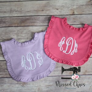 May include: Two ruffled bibs with embroidered initials. The bibs are in pink and lavender colors. The initials are in white thread.
