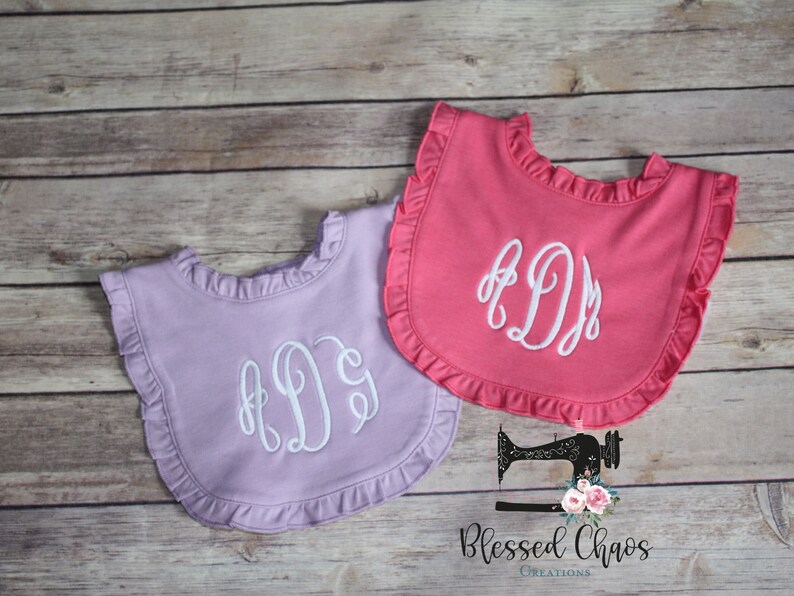 May include: Two ruffled bibs with embroidered initials. The bibs are in pink and lavender colors. The initials are in white thread.