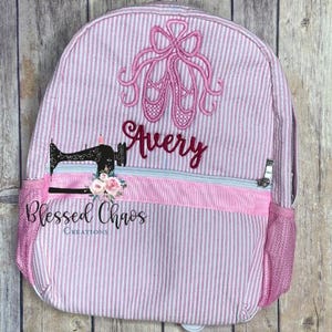May include: Pink and white striped backpack with a pink embroidered ballerina shoe design and the name Avery. The backpack has a pink zippered pocket and a mesh side pocket. The text "Blessed Chaos Creations" is embroidered on the bottom of the backpack.