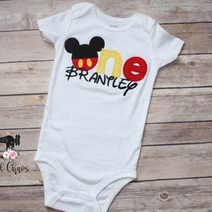 Peut inclure: Combinaison blanche pour b&eacute;b&eacute; avec une t&ecirc;te de Mickey Mouse, un 'un' rouge et jaune et le nom 'Branpley' brod&eacute; en noir sur le devant.