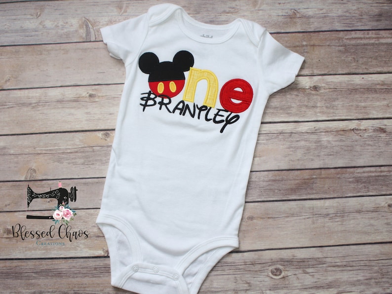 Peut inclure: Combinaison blanche pour b&eacute;b&eacute; avec une t&ecirc;te de Mickey Mouse, un 'un' rouge et jaune et le nom 'Branpley' brod&eacute; en noir sur le devant.