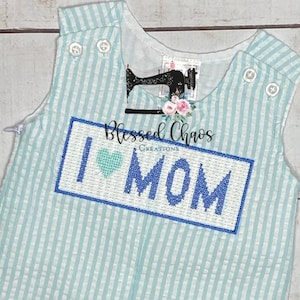 May include: A light blue and white striped baby onesie with a blue embroidered rectangle that says "I love MOM" with a blue heart in the middle. The onesie has a small white tag that says "Blessed Chaos Creations".