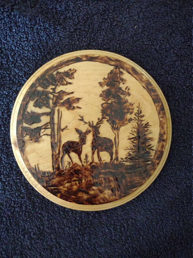 K&ouml;nnte beinhalten: Runde Holzplakette mit einem Brandmalerei-Design von zwei Hirschen in einer Waldszene. Das Kunstwerk ist in das helle Holz gebrannt, mit dunkelbraunen Details. Die Plakette hat einen nat&uuml;rlichen Holzrand.