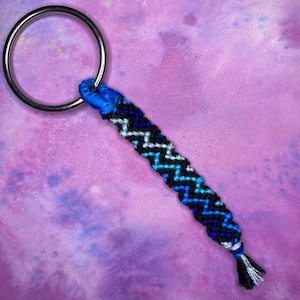 May include: A blue, black and white braided keyring with a silver key ring. The keyring is made with a chevron pattern.