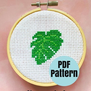Op de afbeelding: Een witte kruissteek borduurring met een groen monstera blad ontwerp. De ring is gemaakt van hout en heeft een metalen klem. De tekst "PDF Pattern" is gedrukt op een blauwe cirkel onder de ring. De tekst "Monstera Leaf" is gedrukt onder de blauwe cirkel.