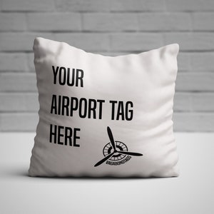 Peut inclure: Un oreiller blanc avec du texte noir qui dit "YOUR AIRPORT TAG HERE" et un logo d'hélice d'avion noir avec le texte "VAGABONDTOURS" en dessous.