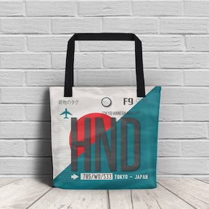 Puede incluir: Una bolsa de tela blanca con asas negras. La bolsa tiene un diseño azul y rojo con el texto "HND" en negro y el texto "TOKYO HANEDA AIRPORT" en negro. La bolsa también tiene el texto "785/W0/533 TOKYO - JAPAN" en negro.