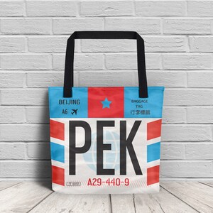 Peut inclure: Un sac fourre-tout blanc avec un design d'étiquette de bagage d'aéroport bleu et rouge. L'étiquette indique "PEK" et "BEIJING" avec une étoile et une icône d'avion. L'étiquette porte également le texte "A29-440-9" et "BAGGAGE TAG 行李牌".